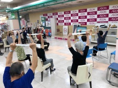 ラジオ体操（写真左）および高岡市が開発した健康体操（写真右）で体を動かす参加者。市の健康体操は、高齢者向きに椅子に座ってもできるようになっている。（写真：2点ともイオンリテール）