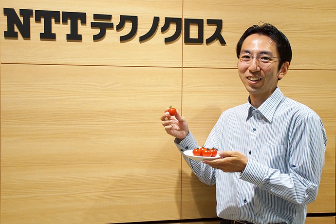 NTTテクノクロスの酒井歩氏。手に持つのはレーザー技術で測定し、かつキャンペーンでも扱っているミニトマトの「あまばんか」