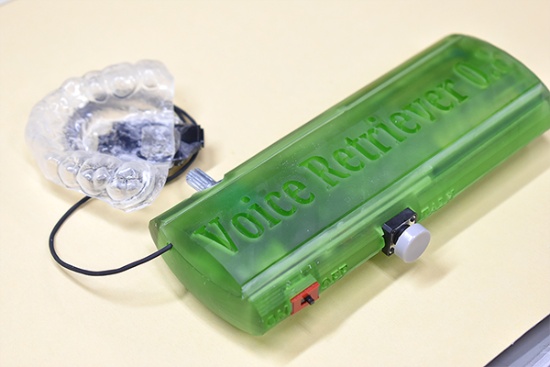 Voice Retrieverの試作機の外観。緑色の外装の機器（手前右）がコントローラー（外部装置）。左のマウスピース型の機器が口内装置で、2つセットで動作する。コントローラーは、電池やしゃべるときに「原音」（詳細は本文で後述）を再生するためのスイッチなどを備える。重量は約100g。重量・形状ともに現在市場に流通している電気式人工喉頭を意識し、持ちやすくすることを目指した。口内装置は口内（上あご）にはめる。スピーカーを搭載しており、ここから声を出すための原音が発せられる。マウスピースは利用者向けに歯科治療の要領で上あごの型を取り、個別に製造する（写真提供：東京医科歯科大学摂食嚥下リハビリテーション学分野、以下同）