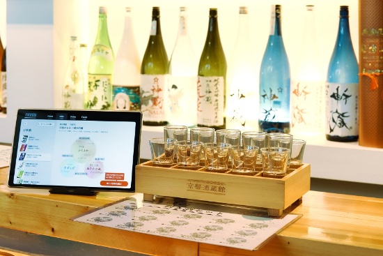 京都酒蔵館のカウンター。中央は「15種類の利き酒セット」の京都北中部バージョン。左のタブレット機の画面に示されたのが日本酒ソムリエAIの「KAORIUM for Sake」（写真撮影：山本真梨子）