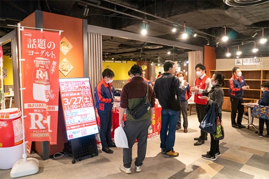 明治クッカーが千葉県松戸市のショッピングモール「キテミテマツド」で行った試飲会および健康イベントの様子。同社が明治特約店として販売している商品の代表例は、「明治プロビオヨーグルトR-1」の宅配専用版。ドリンクタイプ(写真左手ののぼりを参照)と食べるタイプの2種類がある。市販製品とは味や舌触りも明確に違うように作られている。訪問営業などを通じてヒットしており、高い機能性が健康を気にする顧客に刺さった結果だ。加えて明治クッカーでは駅前でのサンプル試飲など独自の取り組みを進め、新たな顧客を獲得しているという(写真提供:明治クッカー)